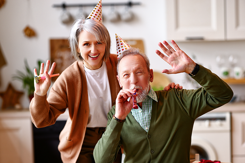 Positive elderly couple celebrating birthday at home, waving at camera. Happy senior man and woman in party hats blowing whistles and smiling, selective focus on people. Holiday celebration concept 